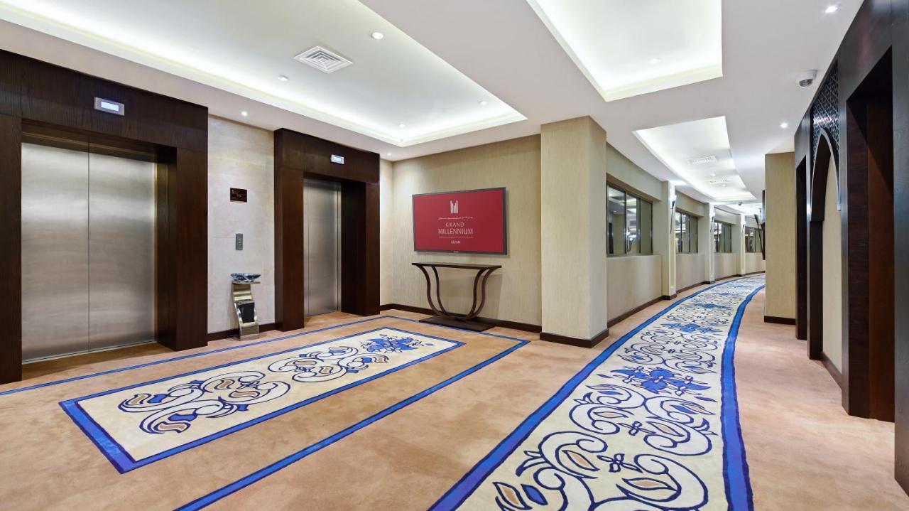 Grand Millennium Gizan Hotel Jizan Exterior photo The photo shows a hotel corridor featuring two elevator doors on the left side. To the right, there is a wall with a red sign, likely indicating the name or logo of the hotel. The floor is adorned with a decorative carpet that has a blue and beige de